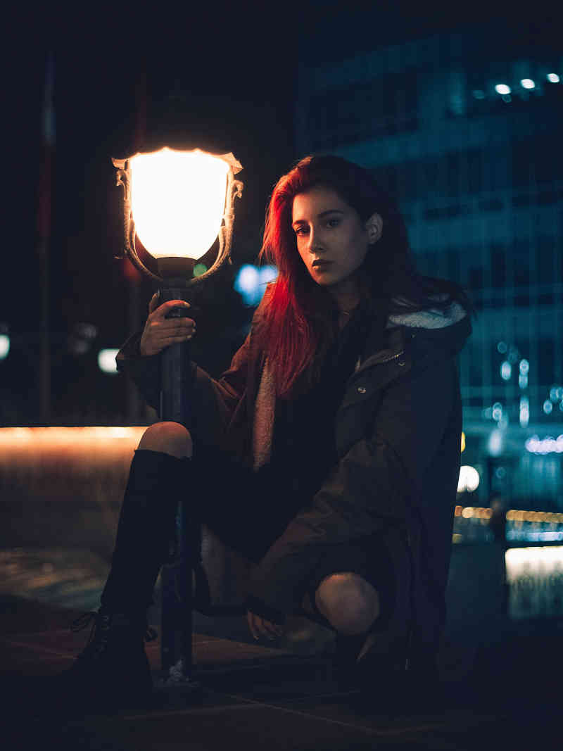 Woman sitting beside, and softly lit by, an ornate outdoor street lamp at night whose upright she holds.